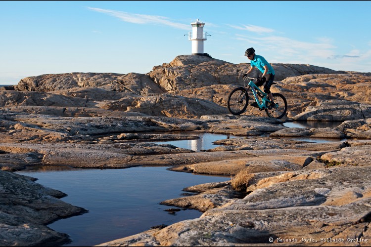 A mountain biker in a blue shirt rides over rocky terrain with a lighthouse in the background. The scene captures a clear sky and small puddles reflecting the surroundings, highlighting the rugged landscape.