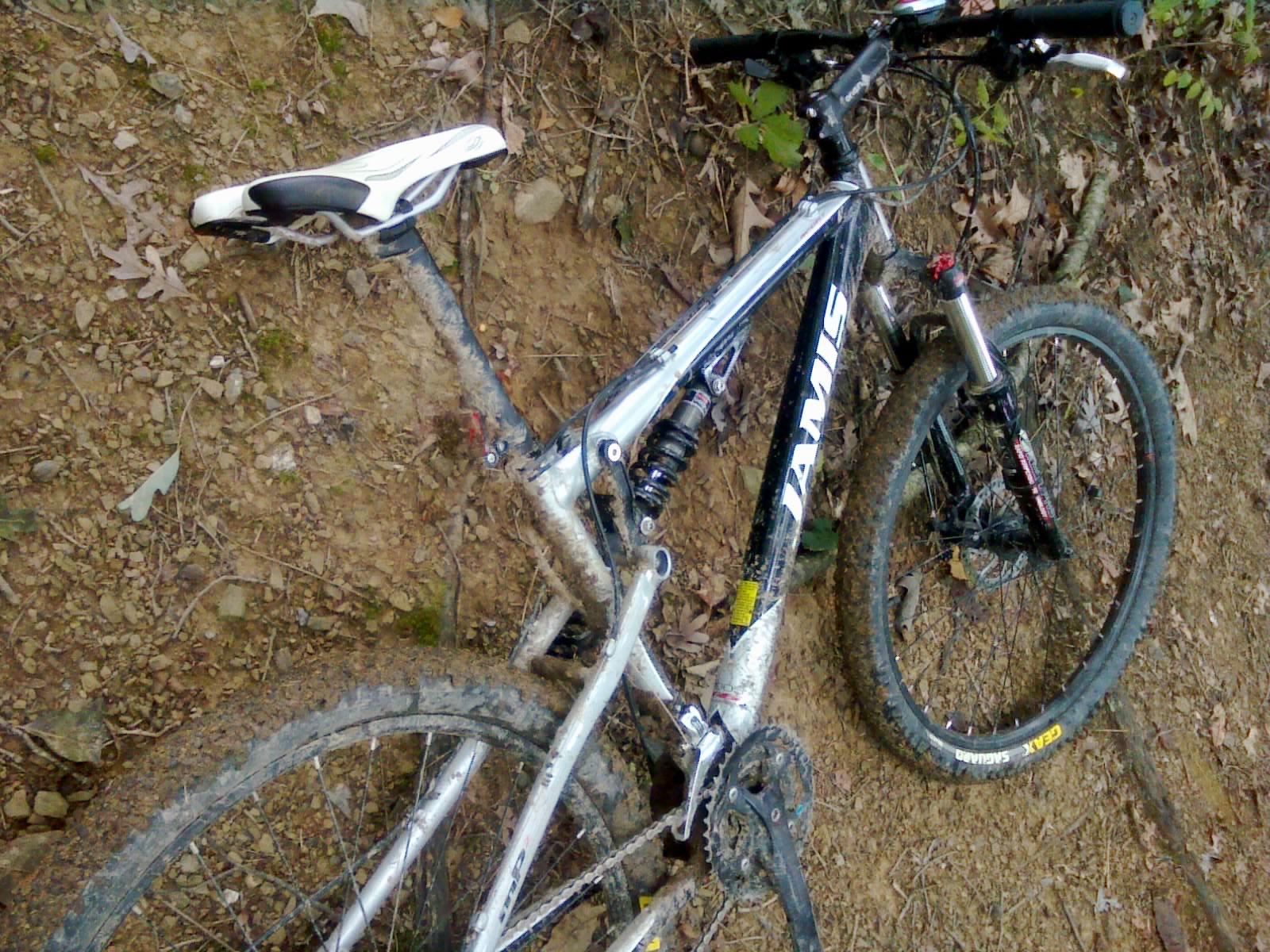 Jamis Dakar XC: A black and silver mountain bike resting on a dirt path, partially obscured by mud and debris. The bike features front suspension, a prominent seat, and rugged tires. Surrounding the bike is a natural landscape with leaves and soil.