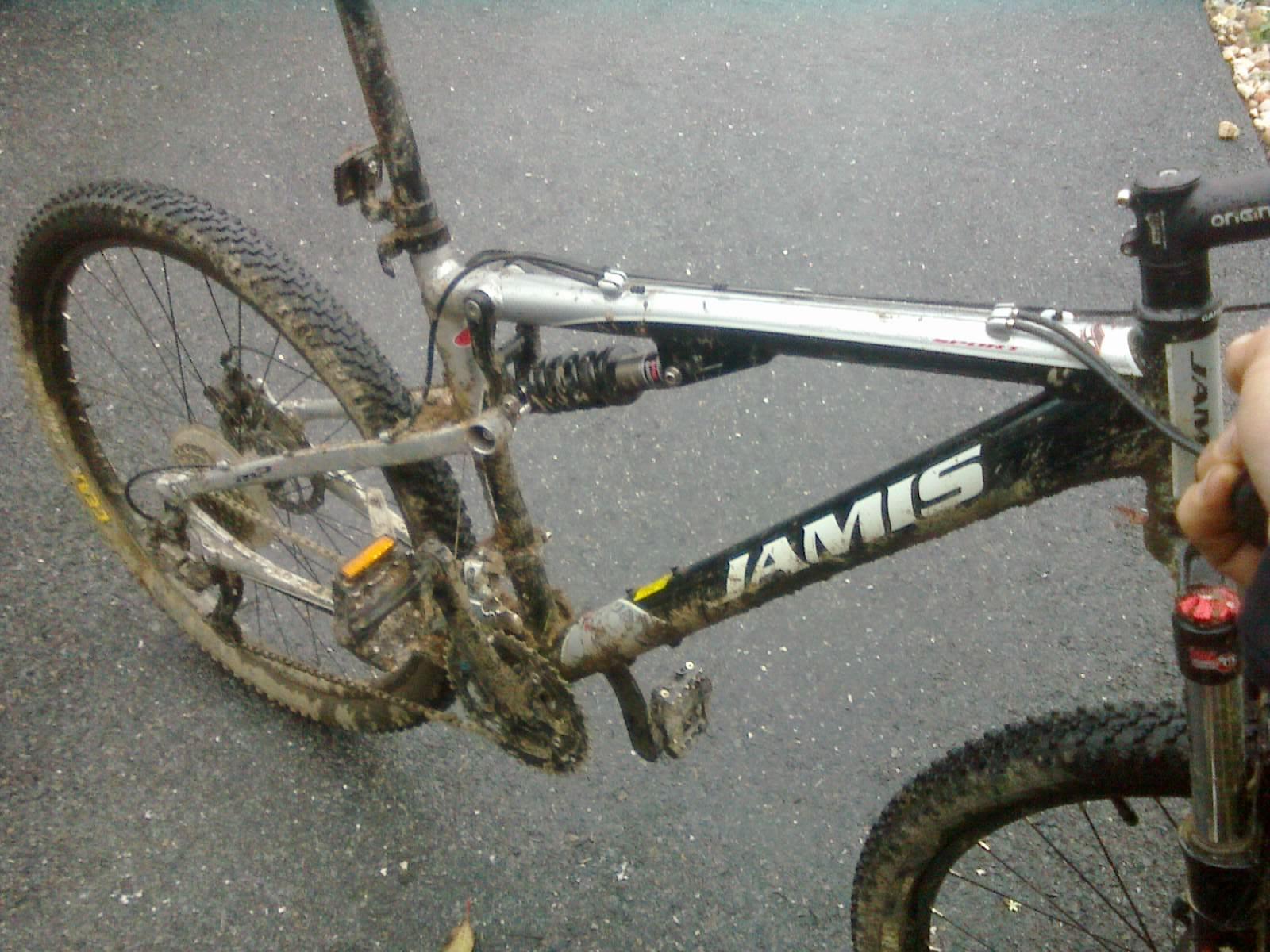 Jamis Dakar XC: A close-up view of a muddy mountain bike, focusing on the frame and drivetrain. The bike features a silver and black design with visible dirt and mud on the wheels and components, indicating recent off-road use. The bike is resting on a gravel surface, with a hand holding the handlebars.