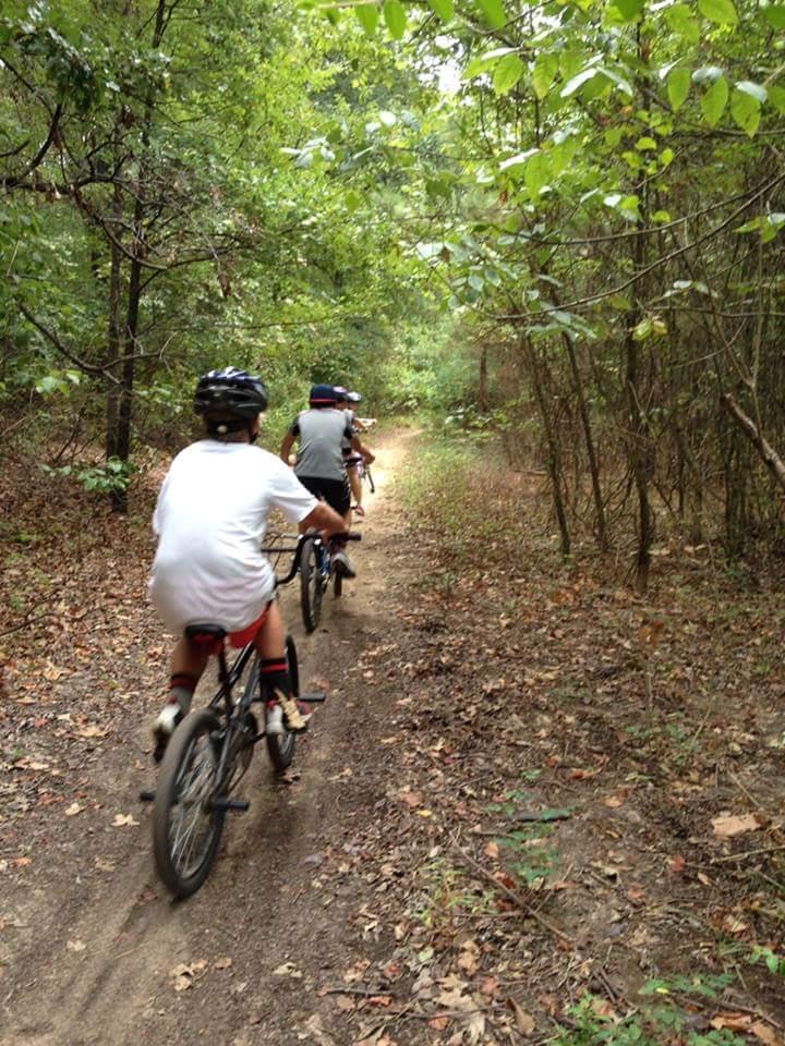 Group of children riding bicycles along a dirt path in a wooded area, surrounded by green trees and foliage. Bringle Lake Mountain Bike Trail System mountain bike trail.