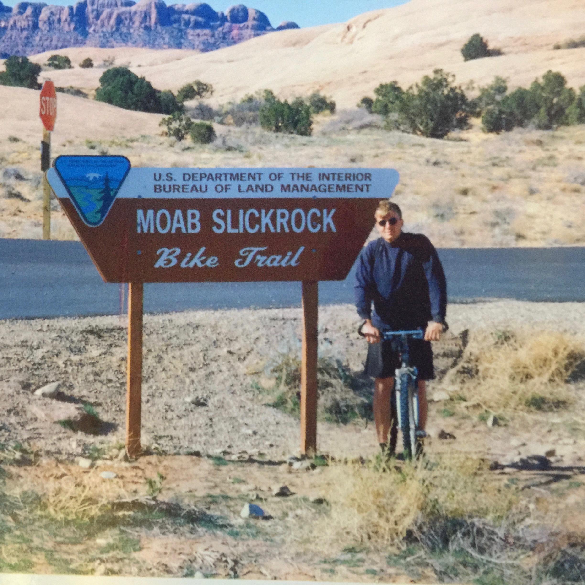 A person standing next to a sign for the Moab Slickrock Bike Trail, holding a mountain bike. The background features arid terrain with rocky formations and vegetation. A stop sign is visible in the area, indicating a road nearby. Slickrock mountain bike trail.