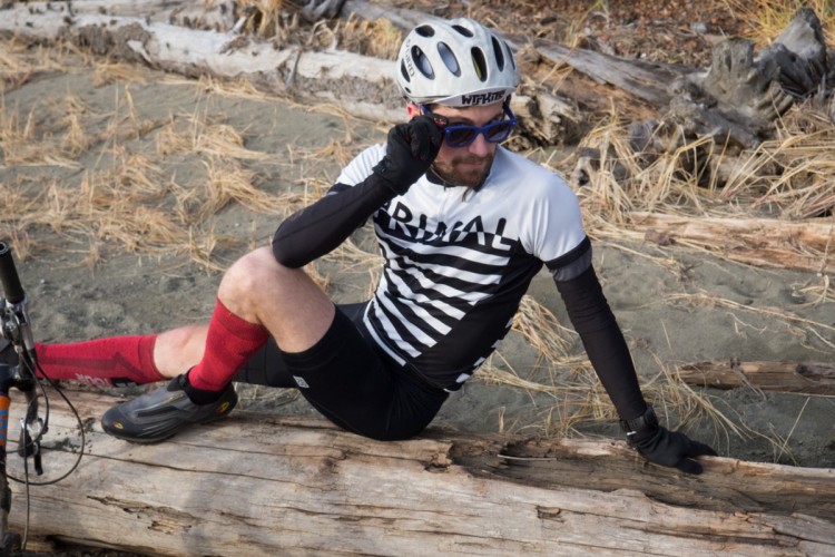 A cyclist wearing a striped jersey and red knee-high socks sits on a log by a sandy area, holding his helmet in one hand and resting the other on his knee. A mountain bike is positioned nearby, and the background features dry grass and fallen logs.
