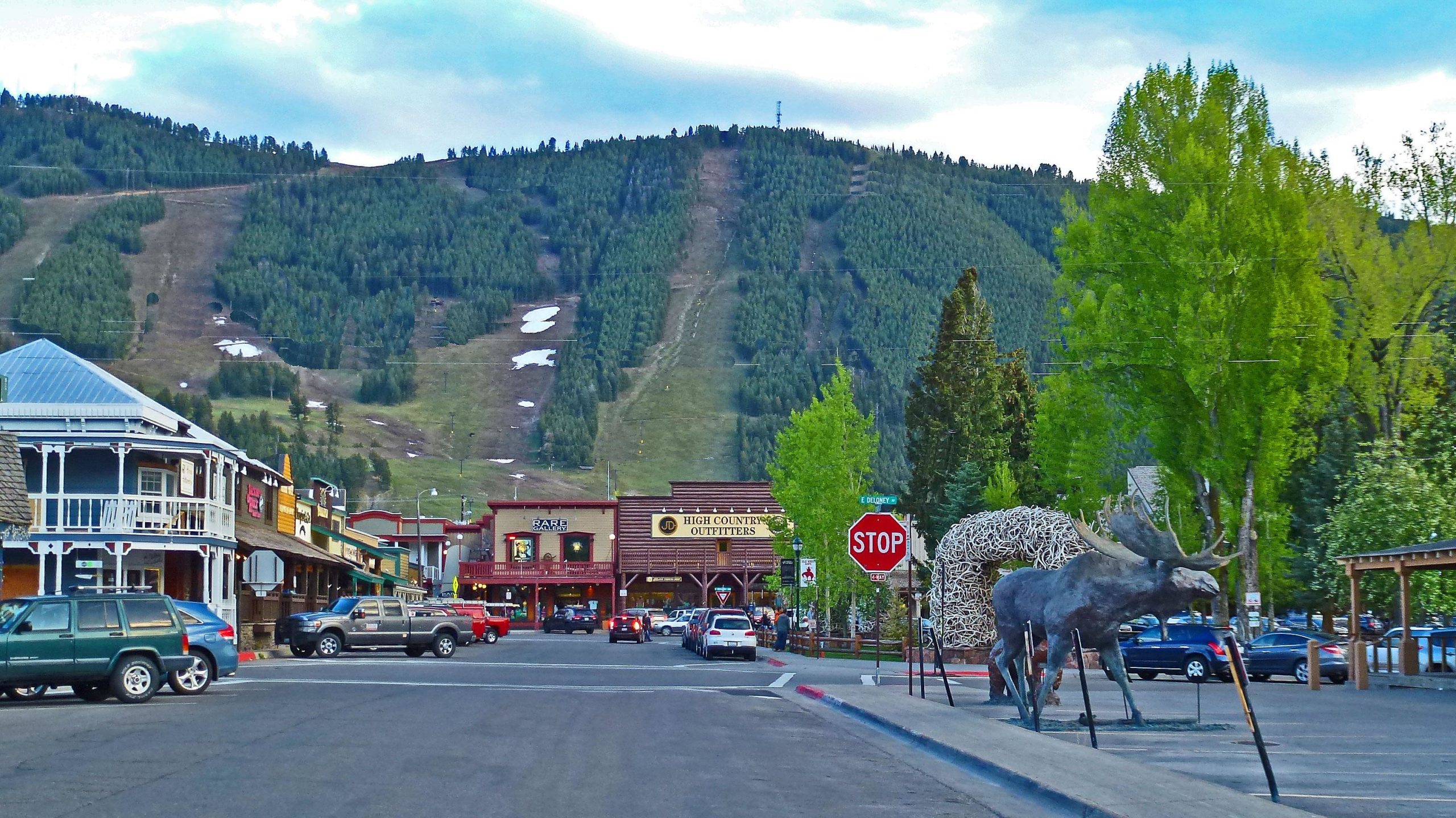 A scenic view of a small town street with wooden buildings, shops, and parked cars. In the foreground, a large, artistic sculpture of a moose made from antlers is visible. In the background, a hillside covered in lush trees under a partly cloudy sky features ski slopes with patches of snow. A stop sign and green trees add to the charm of the location. Snow King Mountain mountain bike trail.