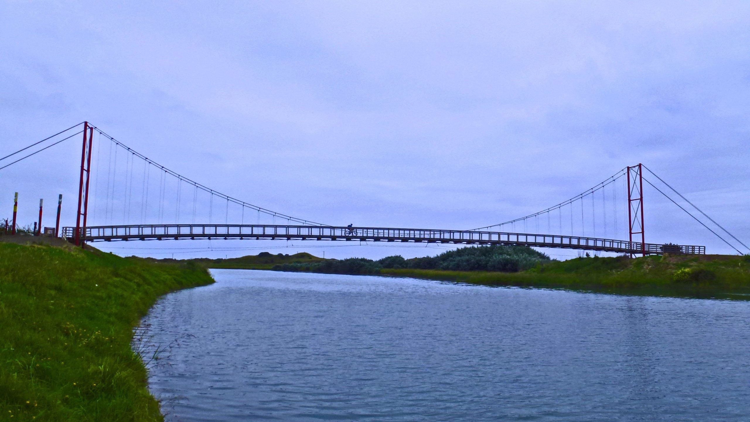 A suspension bridge spans a calm river, with a cyclist visible on the bridge. The scene is set against a backdrop of overcast skies and green grass lining the riverbanks. Dunes Trail mountain bike trail.