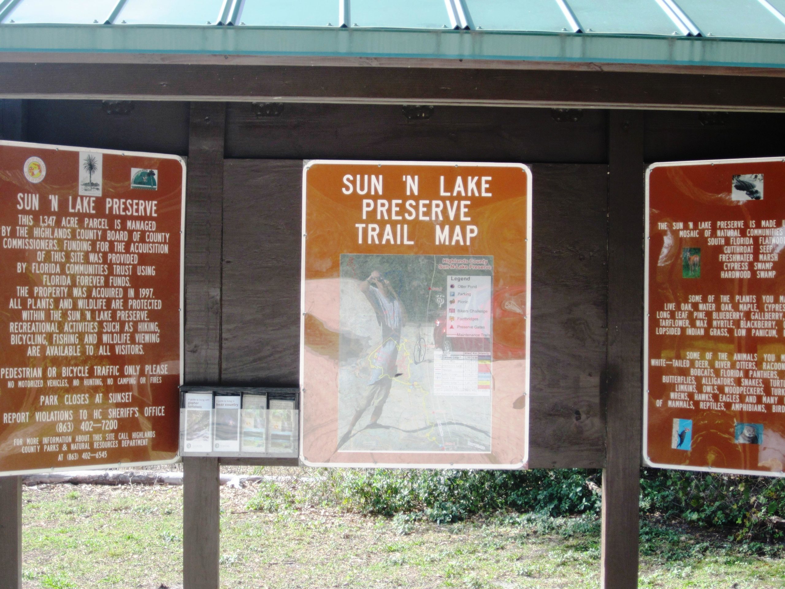 Alt text: A wooden information kiosk at Sun 'N Lake Preserve displaying a trail map and informational signs about the preserve, including details on its management, protected wildlife, and recreational activities available such as hiking and biking. The kiosk features a green roof and is surrounded by lush vegetation. The Preserve mountain bike trail.