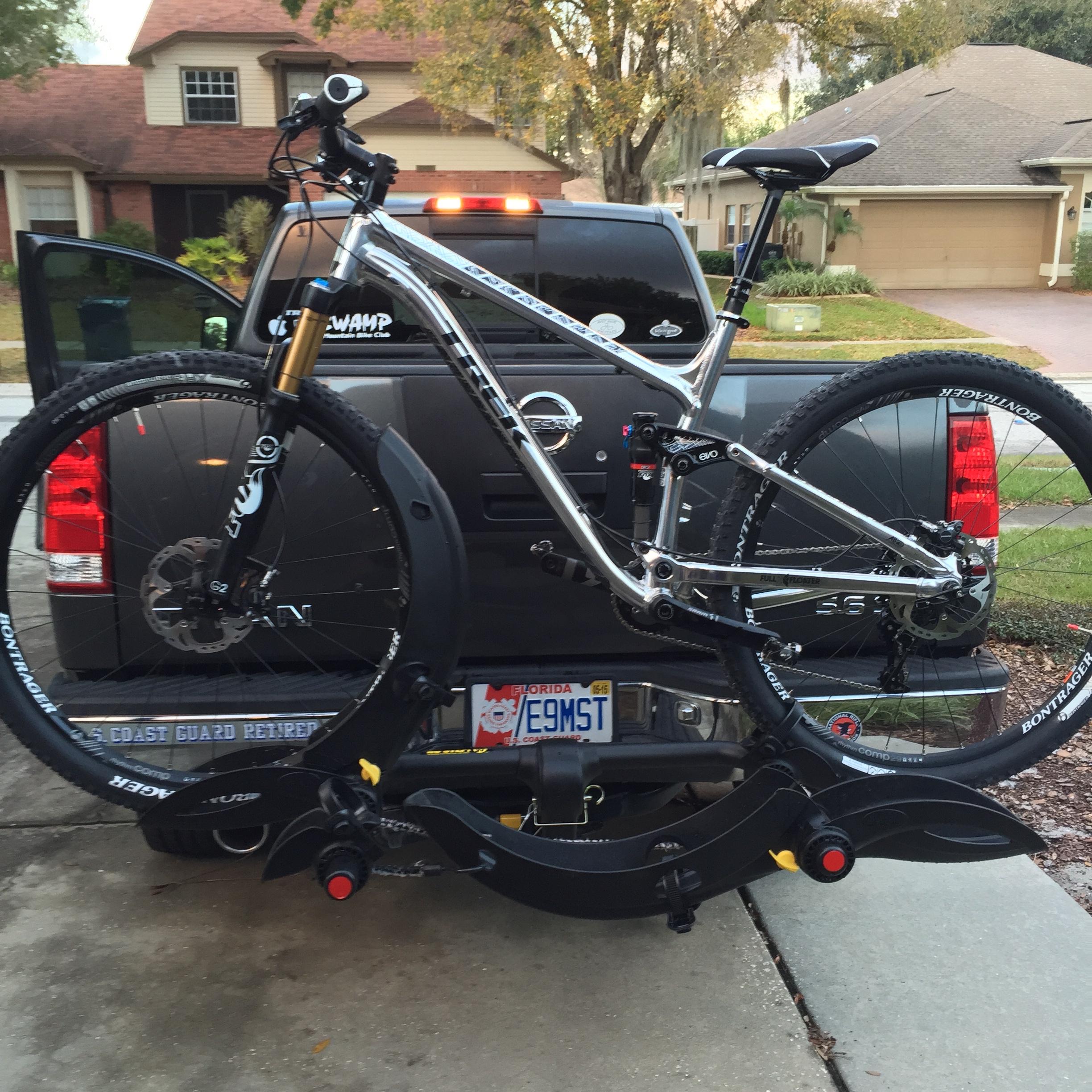 Trek Fuel EX 9 29: A silver mountain bike mounted on a bike rack attached to the back of a pickup truck, with the truck's tailgate open. The background features a suburban neighborhood with houses and trees.