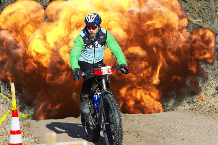 A cyclist in a green and blue jacket rides a mountain bike along a dirt trail, with an explosion of orange and brown smoke in the background. Traffic cones are visible near the path, suggesting a race or event setting.