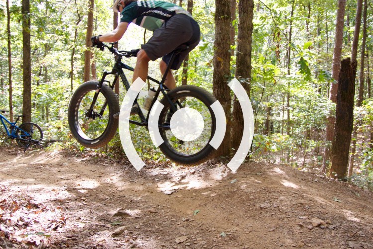 A mountain biker catching air while jumping off a dirt ramp in a wooded trail. A second bike is visible in the background, partially obscured by trees. The image captures a dynamic moment in off-road cycling, surrounded by lush greenery.