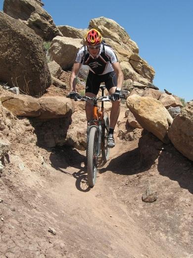 Santa Cruz Superlight: A mountain biker navigating a rocky trail surrounded by large boulders under a clear blue sky. The cyclist is wearing a helmet and cycling attire, focused on maneuvering the terrain.