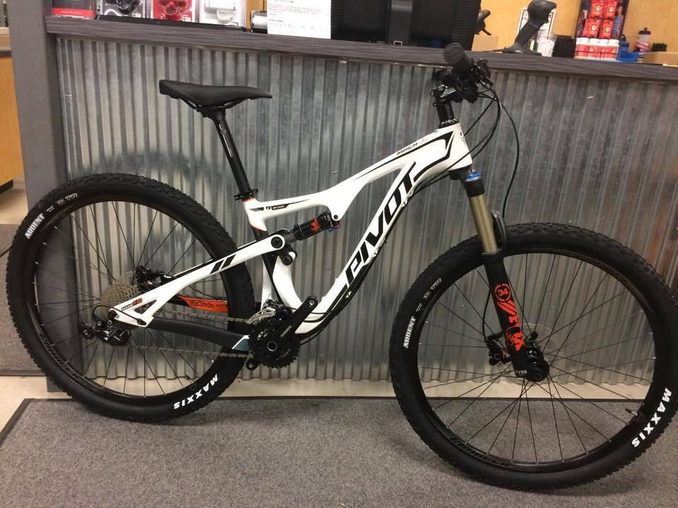 Pivot Trail 429: A stylish mountain bike with a white and black frame is displayed in a bike shop. The bike features thick tires with "Maxxis" branding, a front suspension fork, and a modern design. The background shows a corrugated metal wall and various bike accessories and equipment on display.