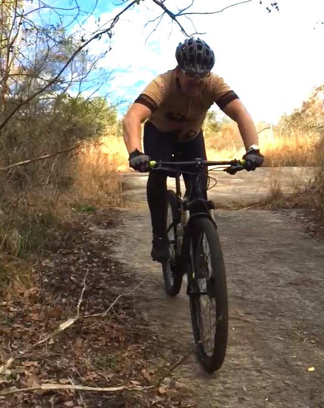 A cyclist wearing a brown jersey and black pants rides a mountain bike on a rocky trail surrounded by grass and trees under a blue sky. Harbins Park mountain bike trail.