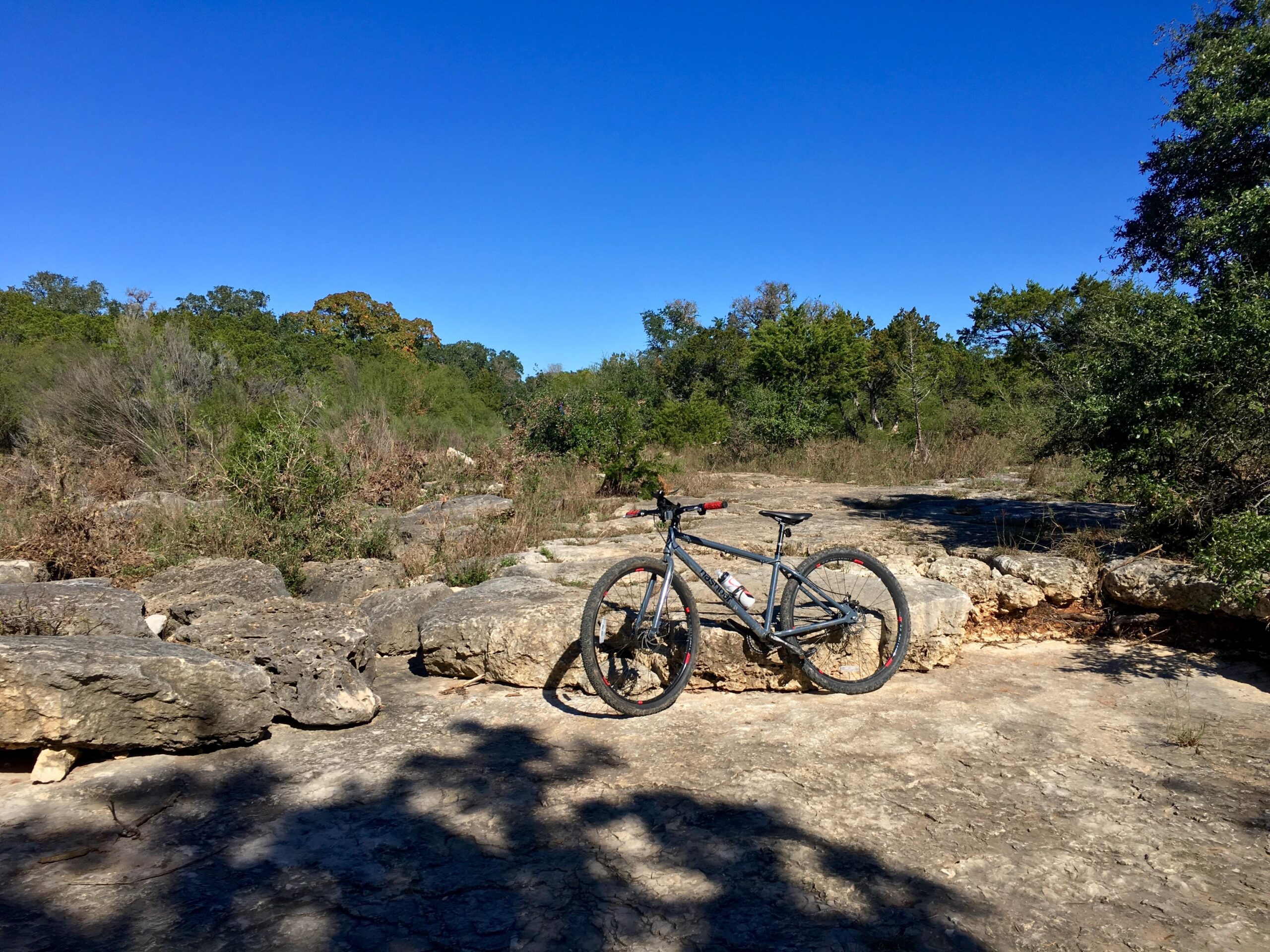 A mountain bike resting on rocky terrain surrounded by lush greenery under a clear blue sky. Salado Creek mountain bike trail.