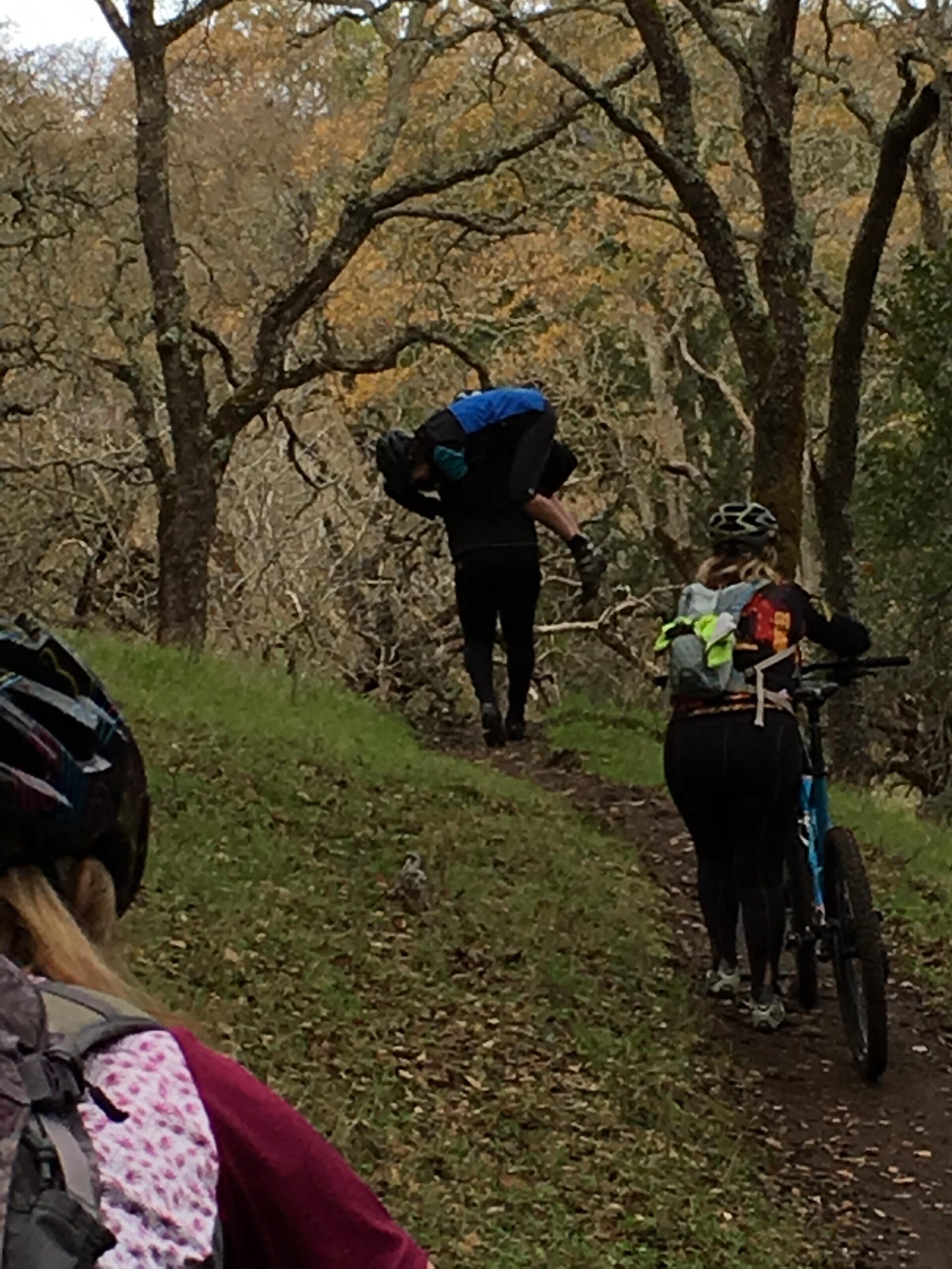A group of mountain bikers navigating a wooded trail. One person is carrying a bike on their shoulder, while another is walking beside a blue mountain bike. The landscape features trees with autumn leaves and a grassy path. Rockville Park mountain bike trail.