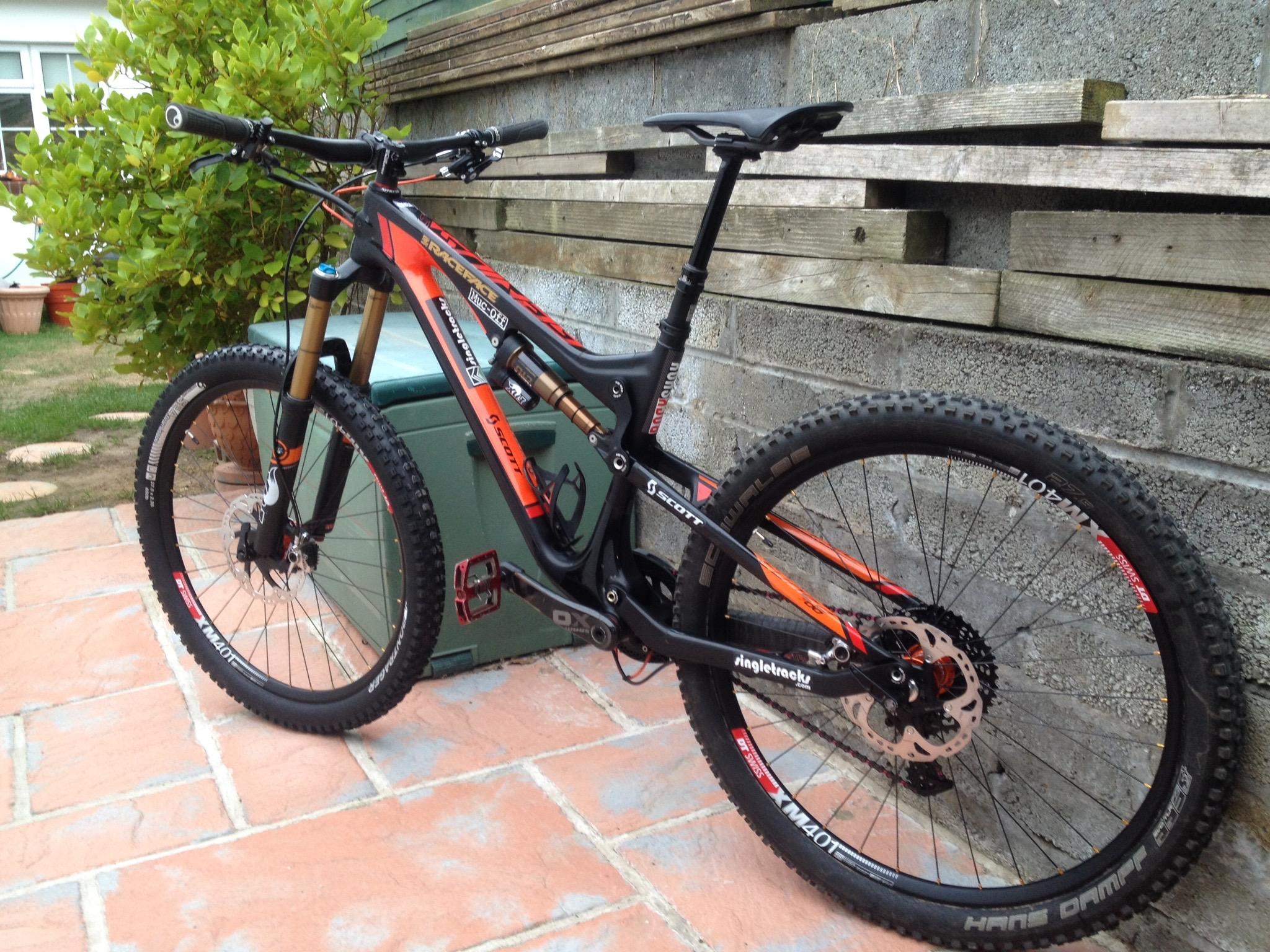 Santa Cruz Bronson: A black and orange mountain bike is positioned against a outdoor wall, showcasing its sturdy frame, thick tires, and suspension forks. The bike is resting on a tiled patio, with greenery visible in the background. The setting suggests a casual outdoor environment, ideal for biking adventures.