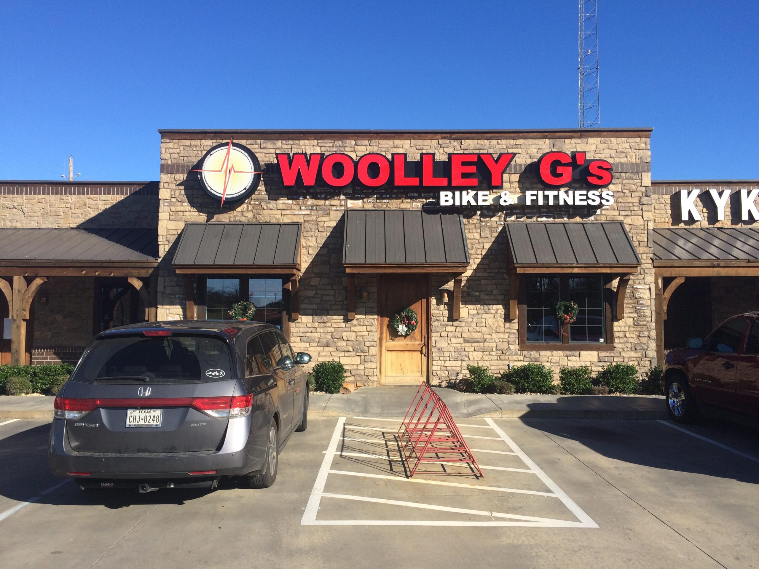 Image of the exterior of Woolley G's Bike & Fitness, a stone building with a sign featuring the business name prominently displayed. In the foreground, there is a parking lot with two vehicles, a grey minivan and a red truck, along with a bike rack. The sky is clear and blue, and the building has holiday wreaths on the windows.