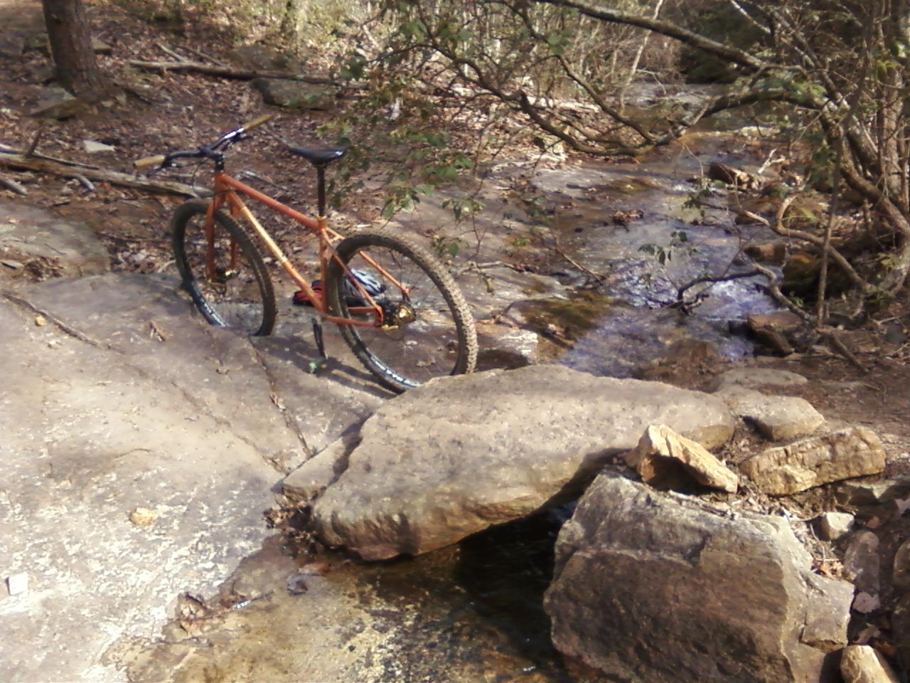 A mountain bike resting on a rocky outcrop near a small stream in a wooded area. The scene features a blend of natural textures, including stones and foliage, creating a serene outdoor setting. Carvin's Cove Trail system mountain bike trail.