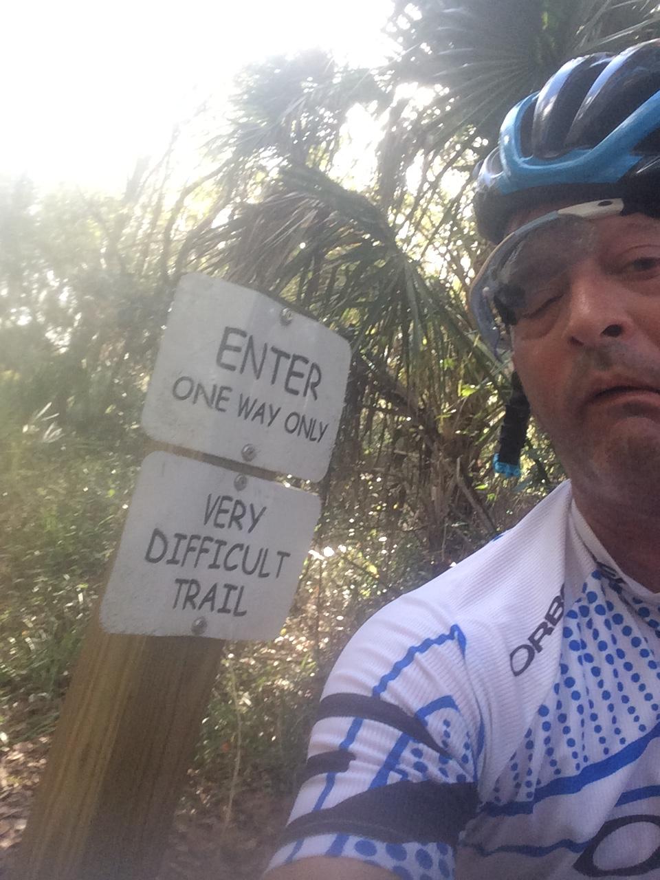 A cyclist wearing a helmet and sunglasses is posing for a selfie next to a trail sign that reads "ENTER ONE WAY ONLY" and "VERY DIFFICULT TRAIL." The background features lush greenery typical of a dense forest. Alafia River State Park mountain bike trail.