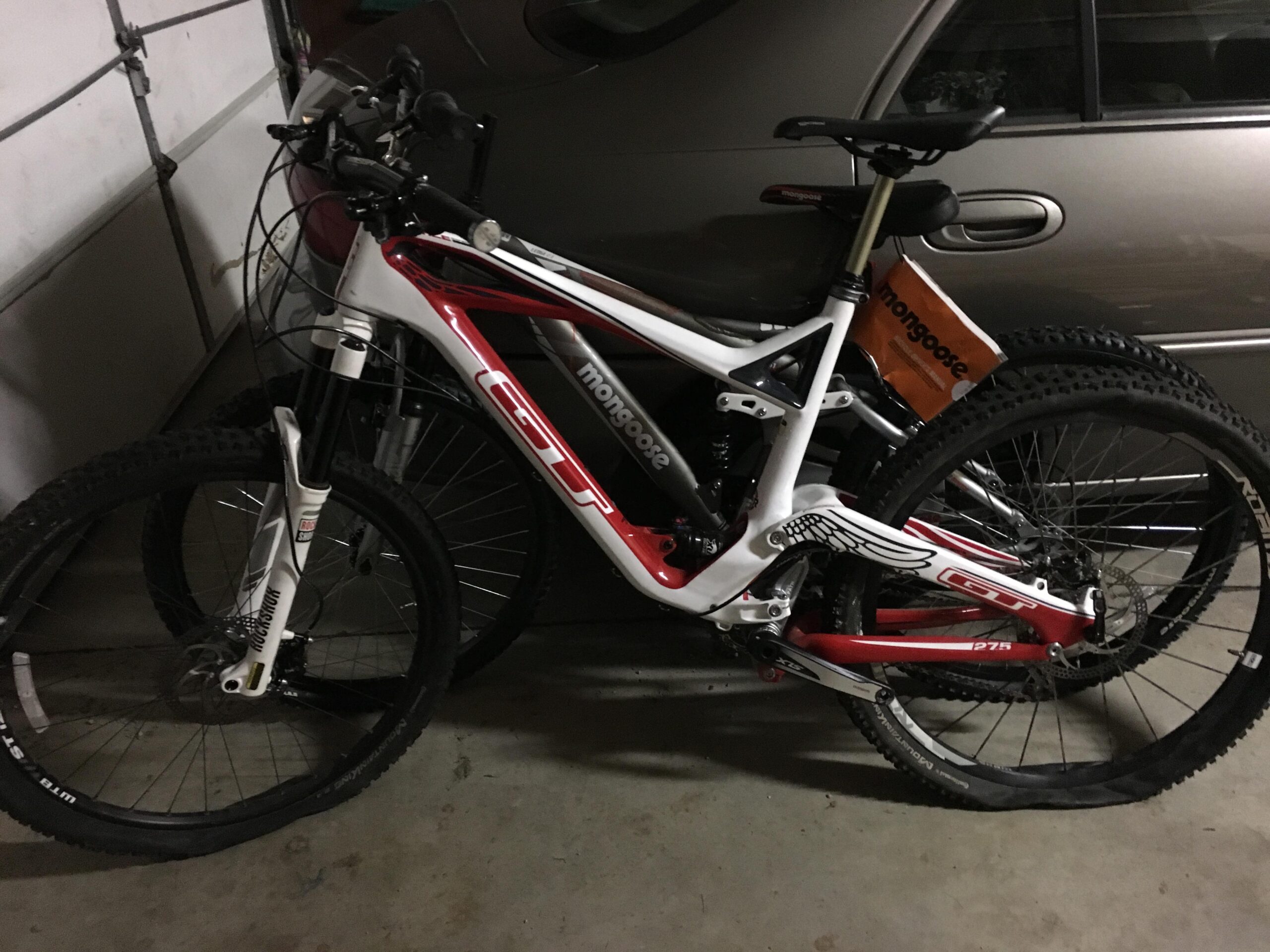 GT Force Carbon Pro: Alt text: Two mountain bikes leaning against each other in a dimly lit garage, with one bike showing a flat tire and a Mongoose brand tag visible on the seat post. A car is parked in the background.