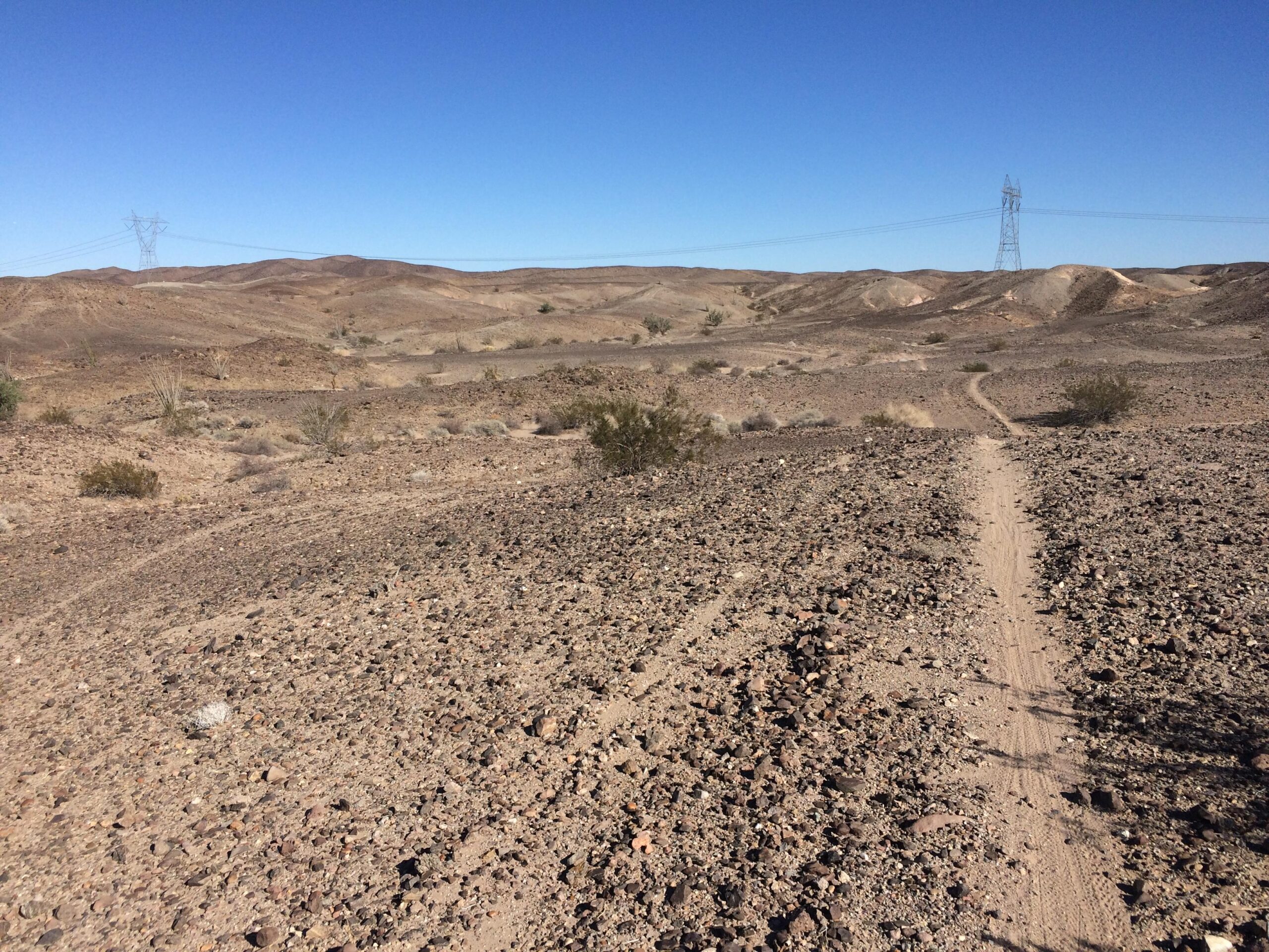 A rocky desert landscape with sparse vegetation, featuring a dirt path winding through the terrain. In the background, rolling hills are visible under a clear blue sky, with utility poles standing along the horizon. Sugarloaf Peak mountain bike trail.