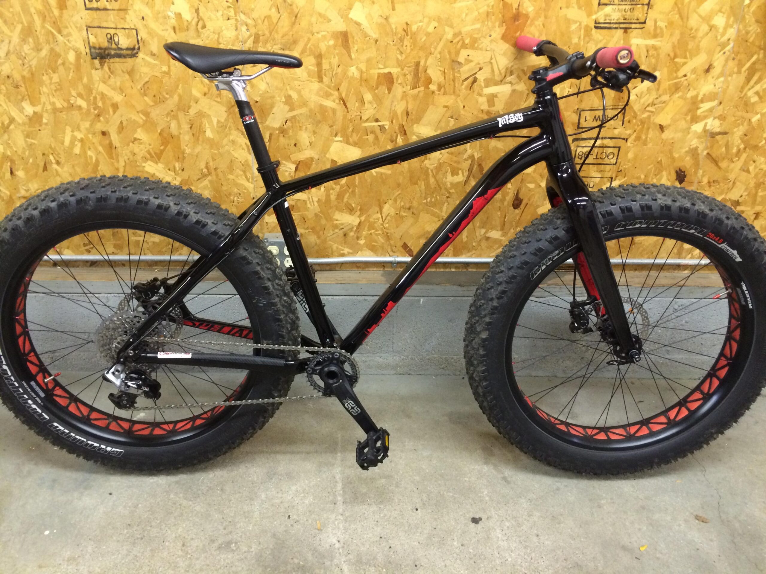 Specialized Fatboy Expert: A black fat tire bike with red accents is positioned in a garage setting. The bike features wide, knobby tires suited for rough terrain, a simple gear setup, and a straight handlebar with rubber grips. The background consists of a wooden wall, adding to the industrial atmosphere of the space.