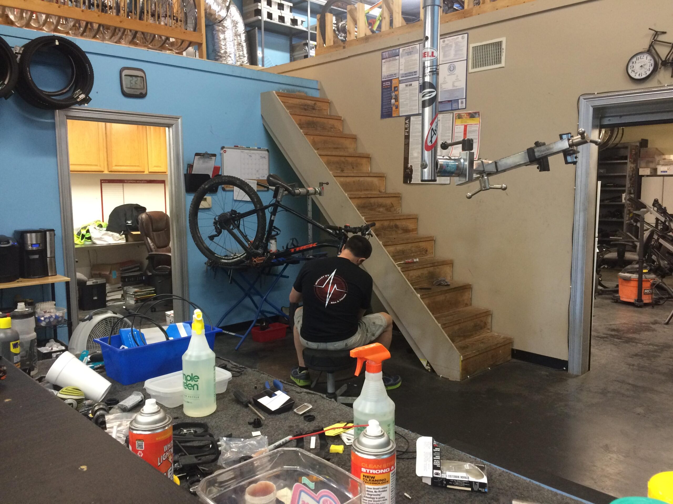 A view of a bike repair shop with a mechanic working on a bicycle. The mechanic is crouched near a workbench, surrounded by tools and bicycle parts. In the background, there is a staircase leading to an upper level, and a bike is mounted on a repair stand. Various cleaning supplies and equipment are scattered around the floor, reflecting a busy workshop environment.