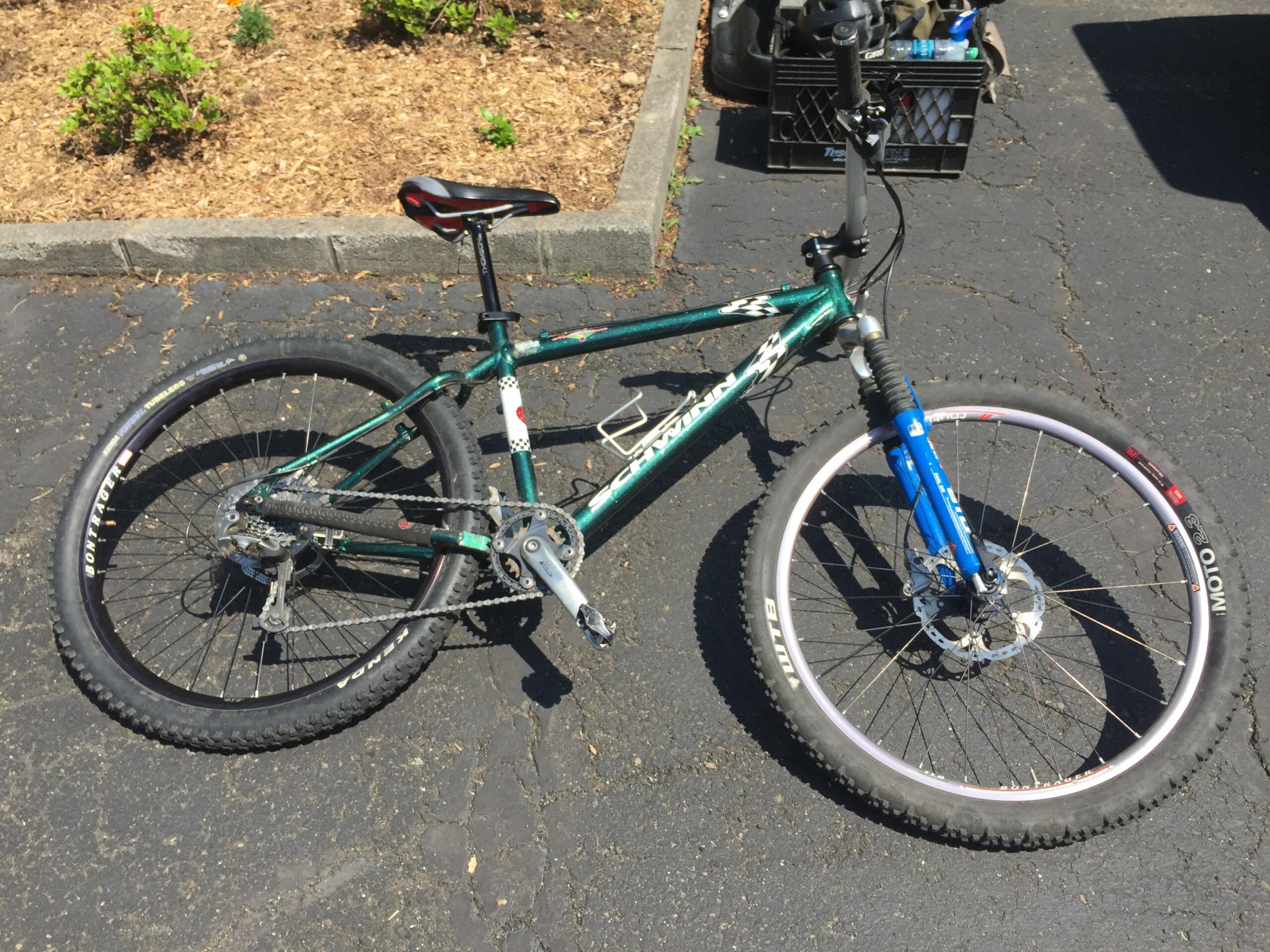 Schwinn Homegrown: A green mountain bike lying on its side on a paved surface, with visible dirt and gravel landscaping in the background. The bike features black tires, a blue front fork, and a multi-speed gear system.