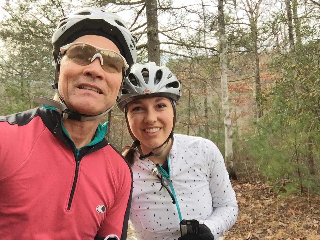 A man and a woman smiling while wearing cycling helmets and clothing, posing for a selfie in a wooded area with trees and foliage in the background. Tsali Recreation Area mountain bike trail.
