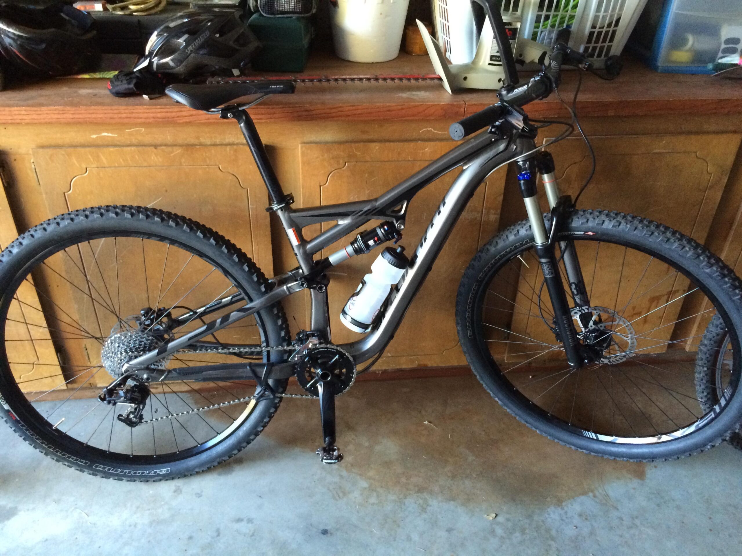 Specialized Camber Comp 29: A mountain bike with a sleek black and gray frame, equipped with thick, rugged tires and a shock-absorbing front suspension, is placed against a wooden workbench. A water bottle is mounted on the frame, and a black helmet is also visible on the bench above. The background features various items, including tools and storage boxes.