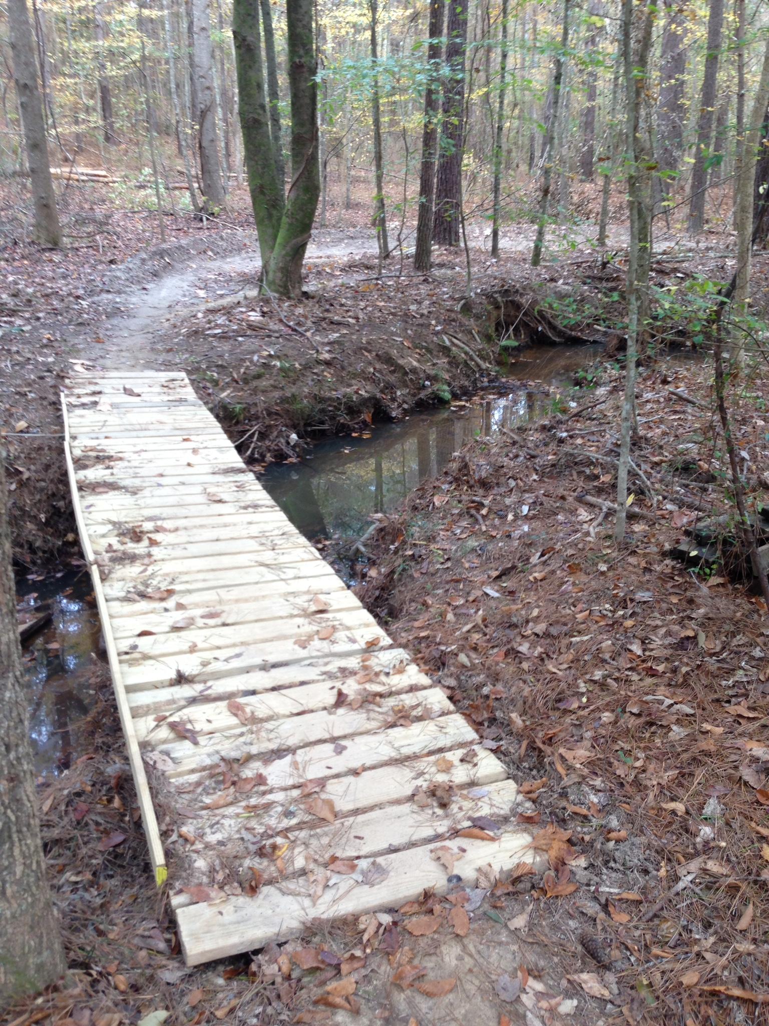 A wooden bridge spans a small creek in a forested area. The surrounding ground is covered with fallen leaves and pine needles, and trees with autumn foliage stand in the background. A winding dirt path leads through the woods, indicating a trail for walking or hiking. Mt. Zion Bike Trails mountain bike trail.