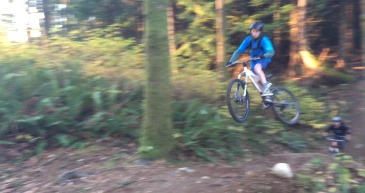 A young person performing a jump on a mountain bike in a wooded area, with trees and plants in the background. Galbraith Mountain mountain bike trail.