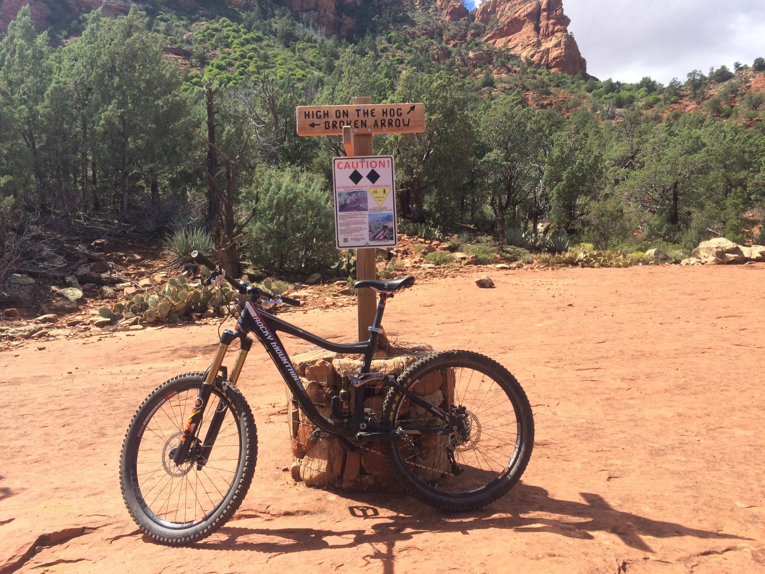 Rocky Mountain Slayer 70: A mountain bike rests beside a trail sign indicating directions for "High on the Hog" and "Broken Arrow." The sign features a caution notice, and the surrounding landscape includes rocky terrain and sparse vegetation typical of a desert environment. Sunlight illuminates the scene, highlighting the reddish tones of the ground and the greenery in the background.