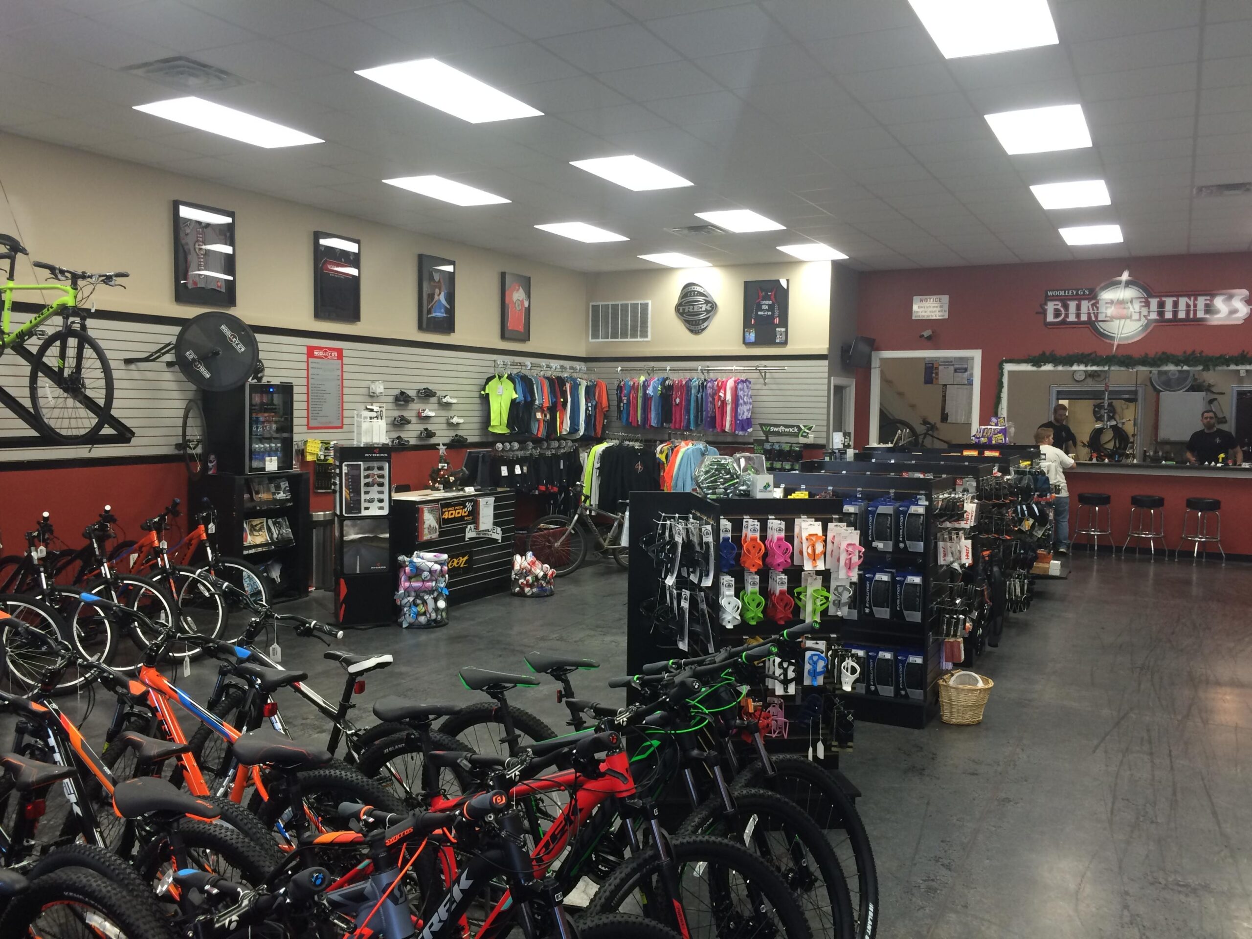 Interior view of a bike shop showcasing a variety of bicycles, including mountain bikes, arranged throughout the space. The store features apparel and accessories displayed on racks along the walls, with merchandise counters and bar stools visible in the background. The environment is well-lit with overhead lighting, and framed sports memorabilia decorates the walls.