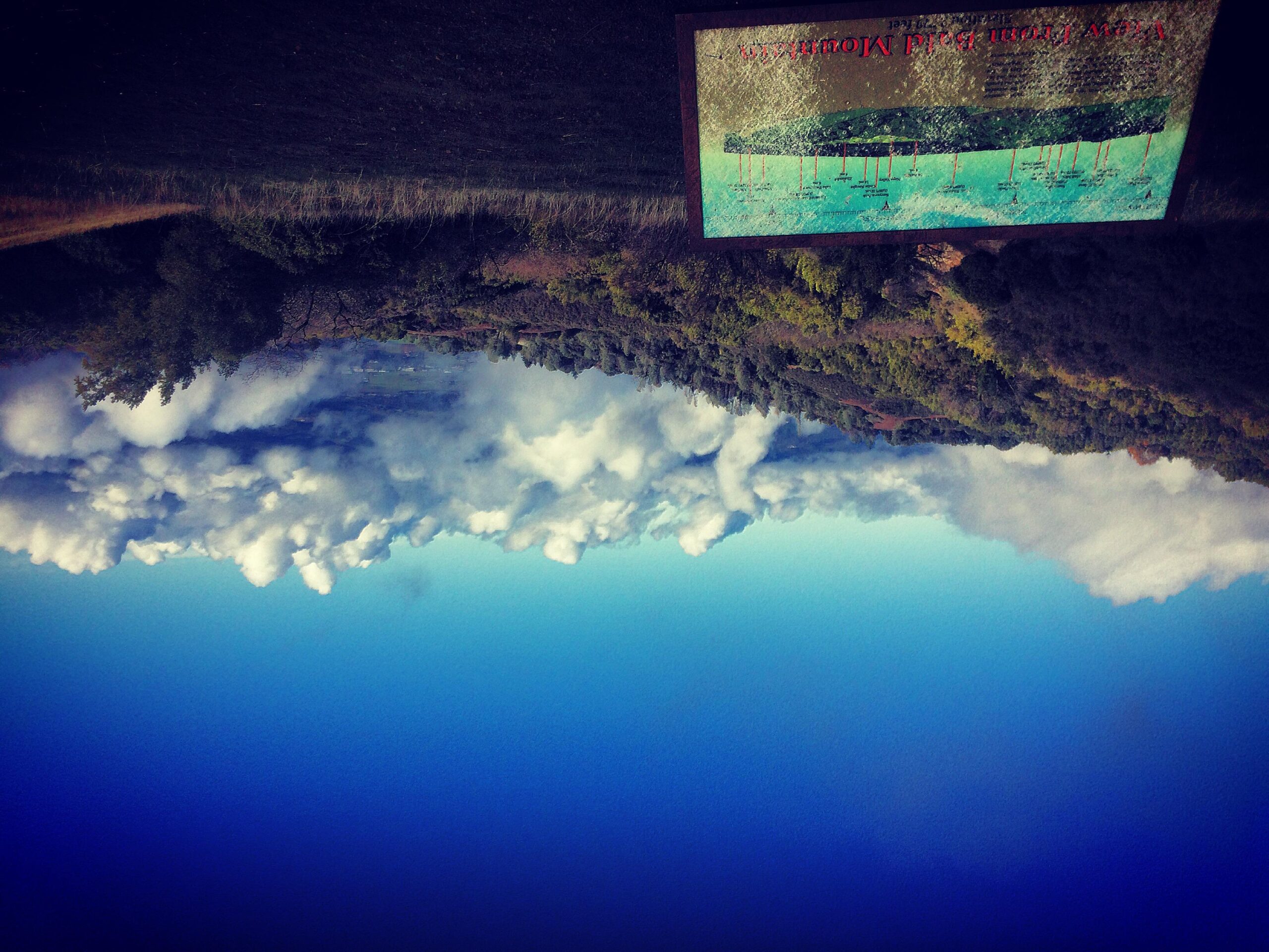 A landscape featuring a vast expanse of blue sky with fluffy white clouds, complemented by a green hillside partially covered with trees. In the foreground, a tilted information sign is visible, displaying a graphic and text related to the view. The overall scene captures a serene natural environment. Sugarloaf Ridge State Park mountain bike trail.