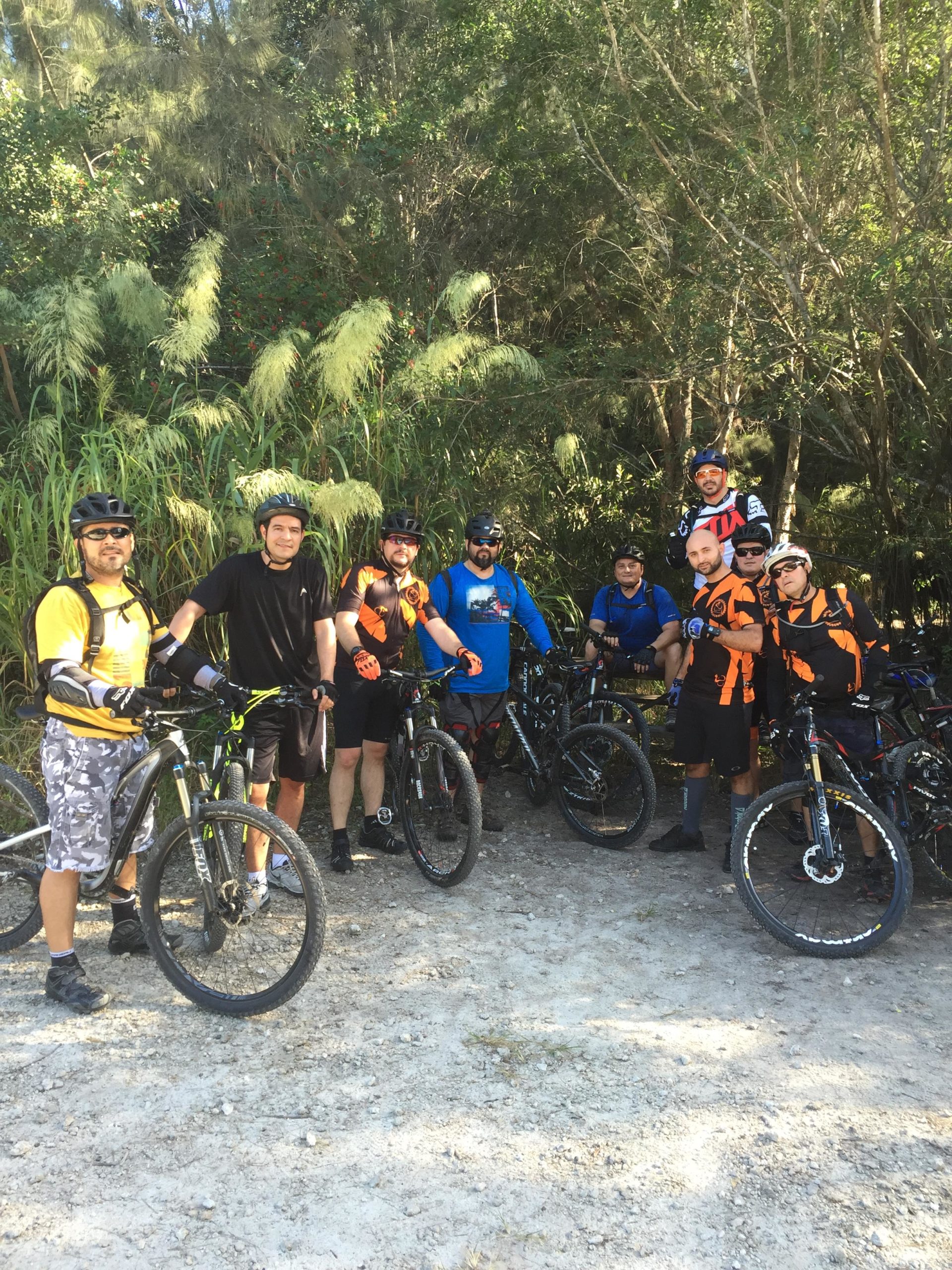 A group of eight mountain bikers posing together on a dirt path surrounded by greenery. They are wearing helmets and various cycling clothing, with their bikes parked beside them. The scene captures a moment before embarking on a ride. Virginia Key North Point mountain bike trail.