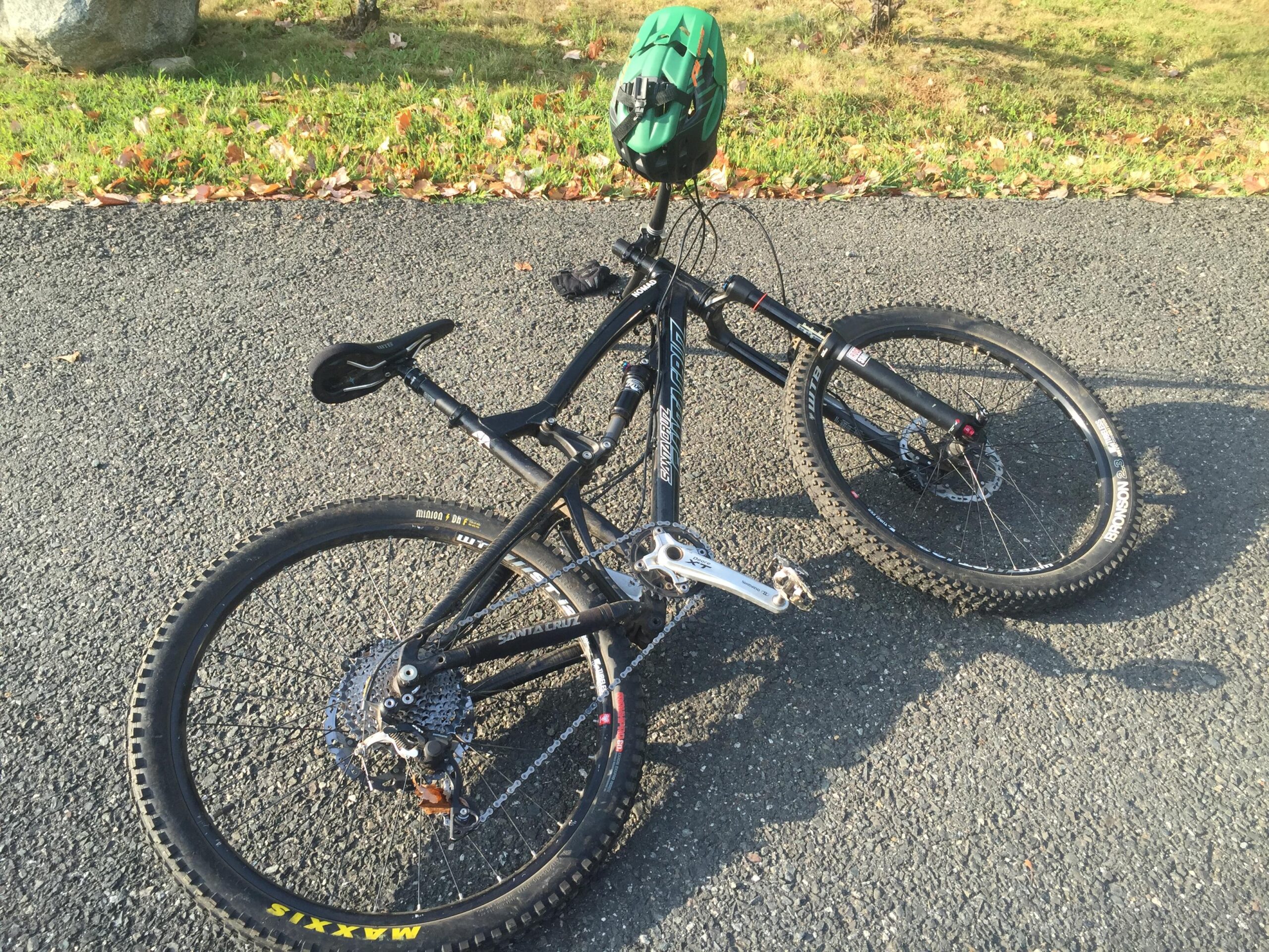 Santa Cruz Nomad: A black mountain bike resting on a gravel path, with a green helmet placed on the handlebars. The bike features thick tires designed for off-road terrain and a visible gear system. In the background, patches of grass and some leaves can be seen.