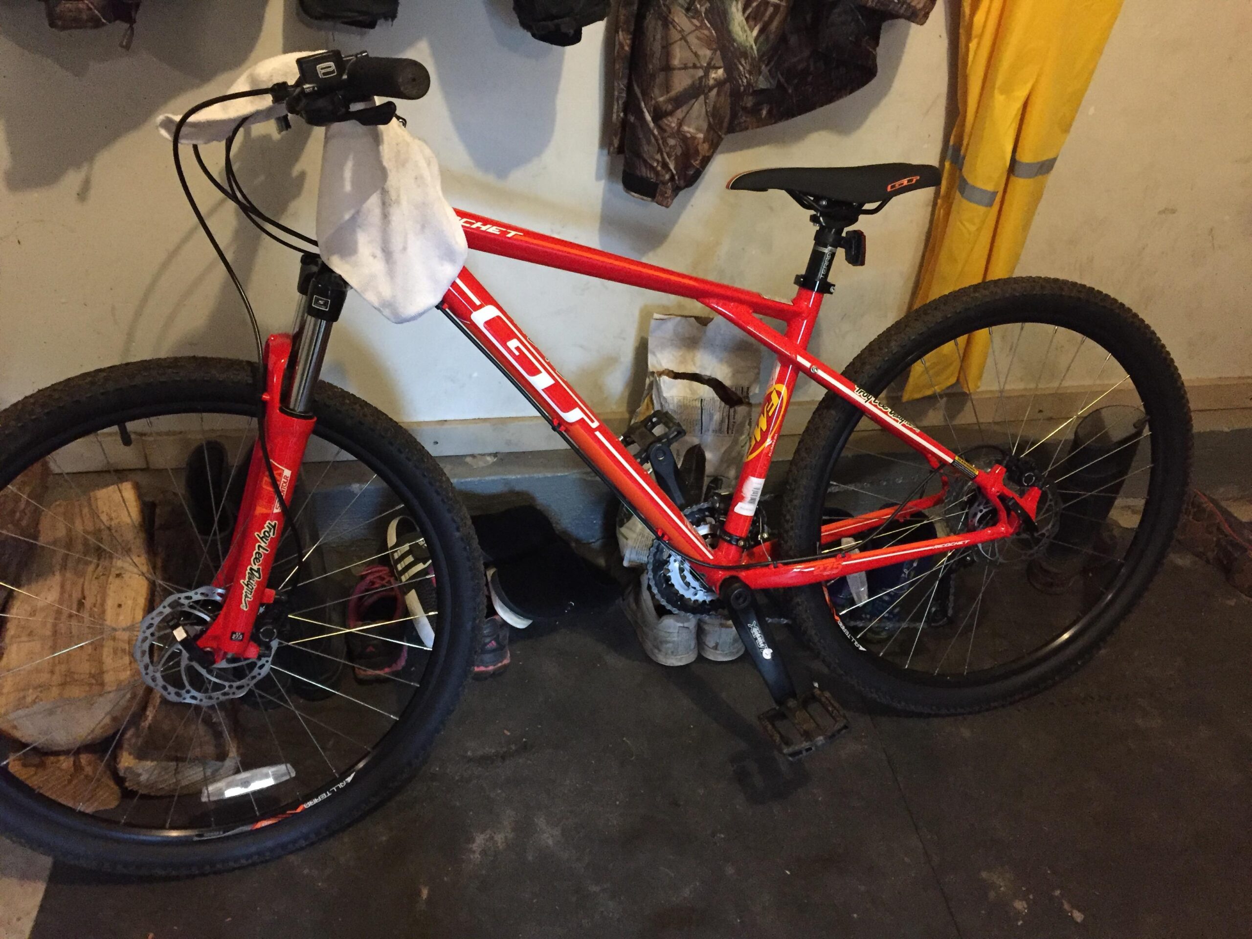 GT Ricochet: A red mountain bike resting on a dark floor, with a towel draped over the handlebars. The bike features thick tires and disc brakes, and is positioned near shoes and wood logs in a cluttered garage setting.