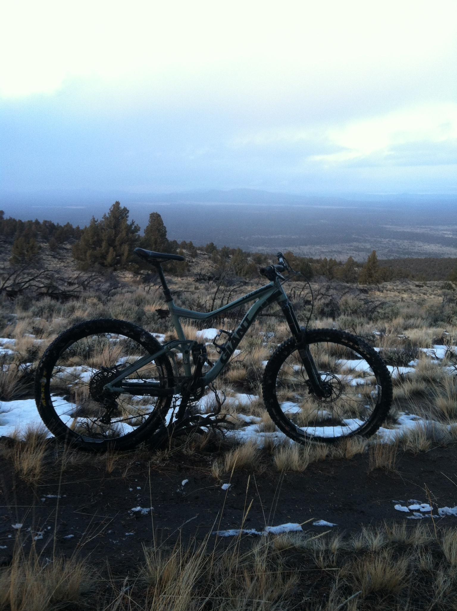 Giant Trance SX 27.5: A mountain bike resting on a dirt path surrounded by sparse grass and patches of snow, with a panoramic view of a valley and mountains in the background under a cloudy sky.