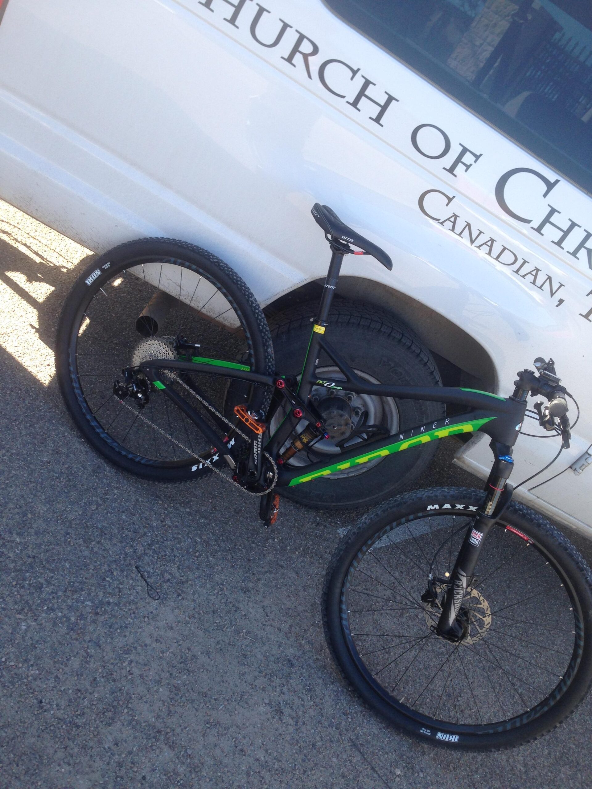A black and green mountain bike parked beside a white vehicle with the text "Church of Christ" visible on the side. The bike features thick tires and a modern frame design, resting on a gravel surface.