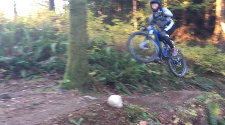 A person in biking gear performing a jump on a mountain bike over a small rock on a dirt trail surrounded by trees and greenery. Galbraith Mountain mountain bike trail.