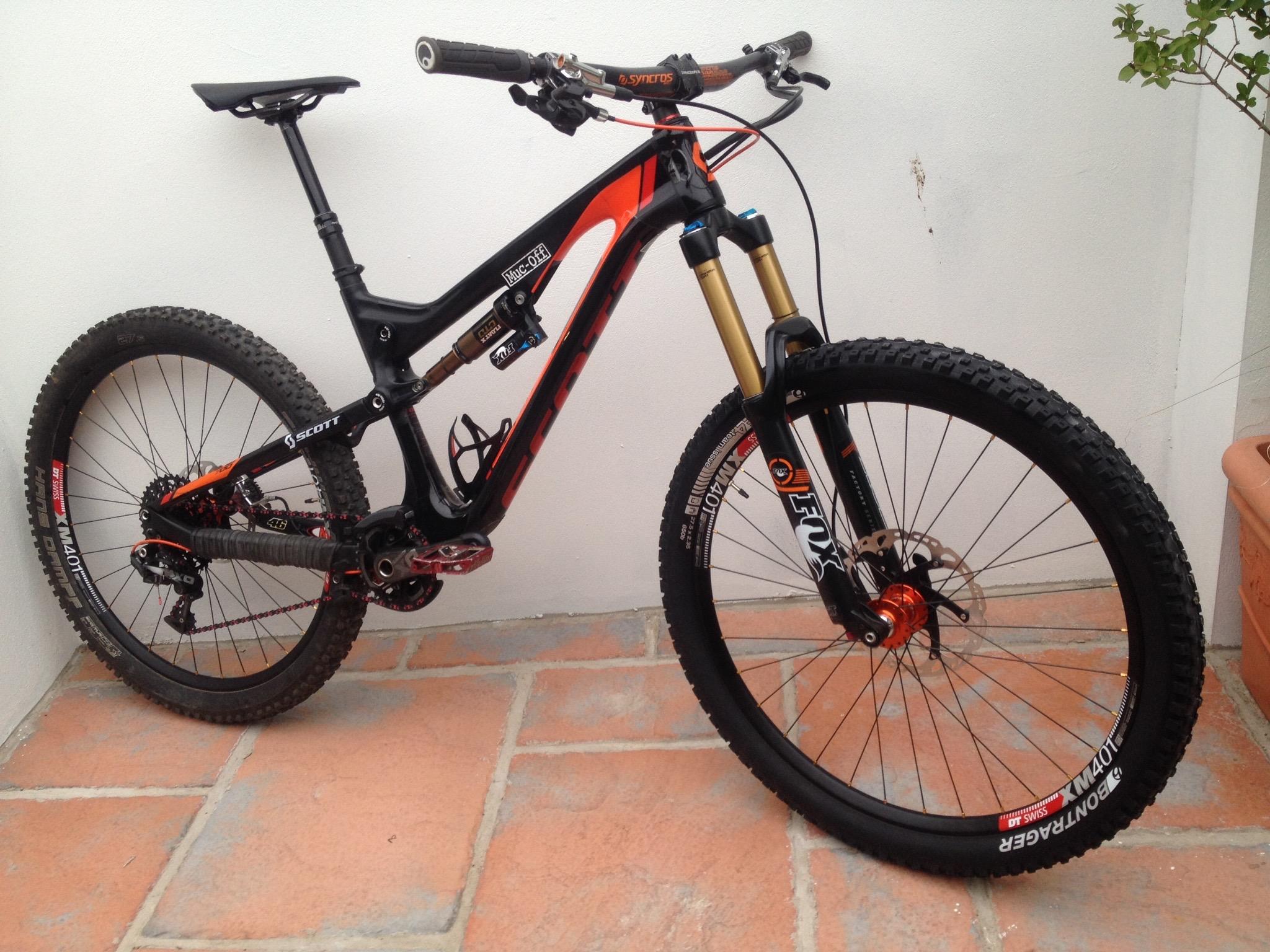 Santa Cruz Bronson: A black and orange mountain bike leaning against a white wall. The bike features a full suspension frame, front forks with gold components, and thick, knobby tires suitable for off-road riding. The setting includes a tiled floor, with a small plant visible in a pot nearby.