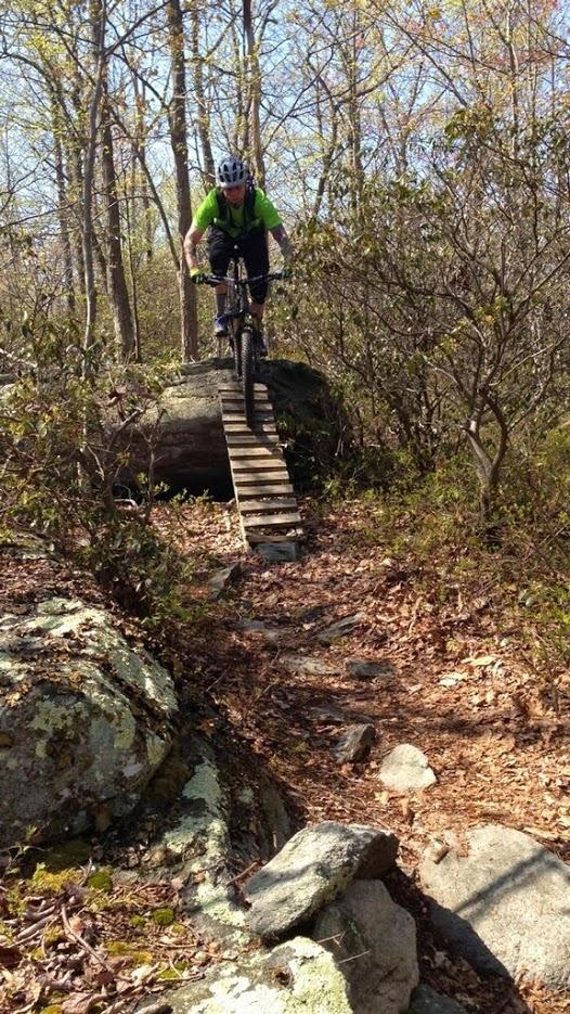 A mountain biker dressed in a bright green shirt and helmet rides over a narrow wooden bridge elevated above rocky terrain. Surrounding the path are trees with budding leaves and scattered dry foliage, creating a natural forest setting. Frederick Watershed mountain bike trail.
