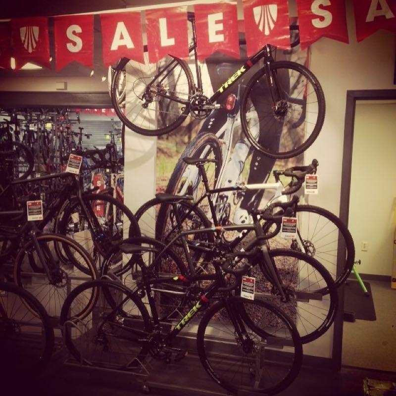 A display of bicycles in a shop featuring a "SALE" banner at the top. Several bikes are hanging on the wall and in a stand, with visible price tags promoting discounts. The backdrop includes a large image of a cyclist riding on a path.