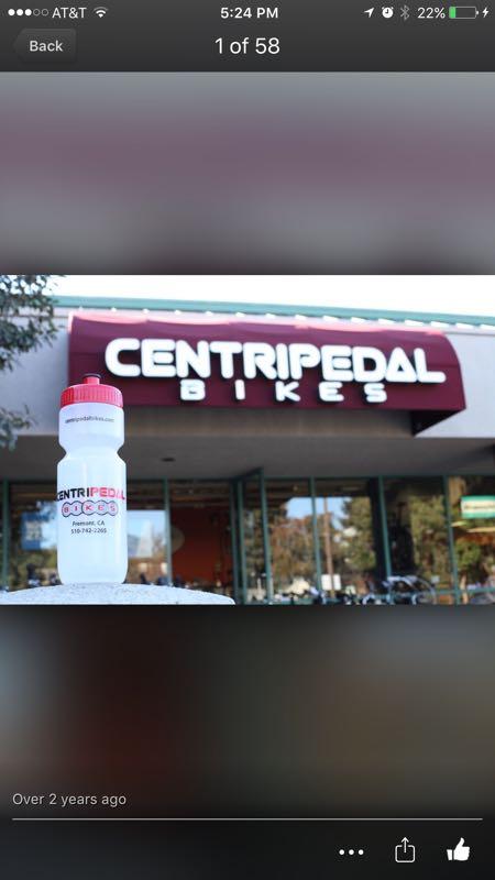 A white sports water bottle with a red cap is placed in front of the storefront of Centripetal Bikes. The shop's sign is prominently displayed above, featuring the name "CENTRIPEDAL" in bold letters, with "BIKES" beneath it. The scene is set outside, with some trees and bicycles visible in the background.