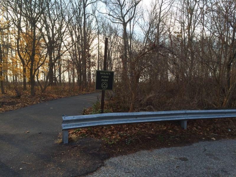 A pathway leading into a wooded area, with a green sign on the left that reads "Wolfe's Pond Park." The scene is partially covered with fallen leaves, and bare trees are visible in the background. A metal guardrail runs along the edge of the paved road. The atmosphere is calm and evokes a sense of nature. Wolfes Pond park mountain bike trail.