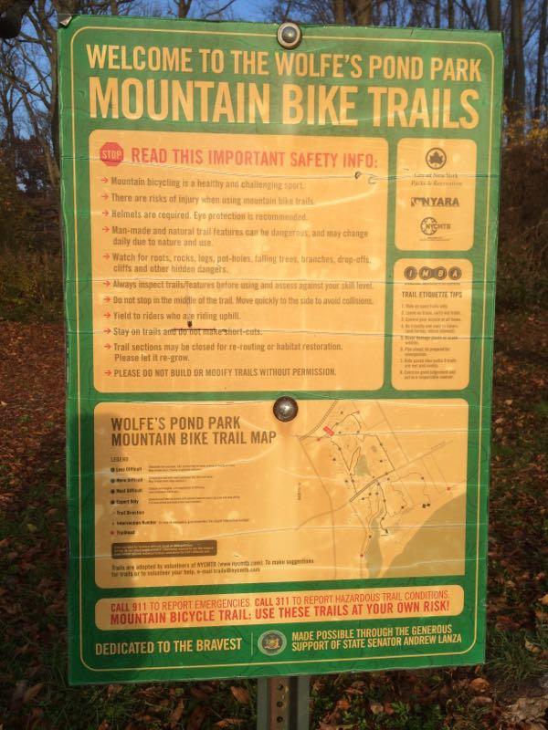 Sign detailing the mountain bike trails at Wolfe's Pond Park, including safety information, trail etiquette tips, and a map of the trails. It highlights the need for helmets, awareness of natural hazards, and encourages responsible trail use. The sign also provides emergency contact details and emphasizes the importance of preserving the trails. Wolfes Pond park mountain bike trail.