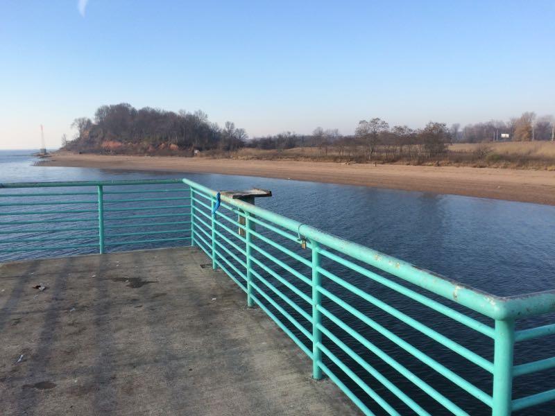 A view from a turquoise railing on a dock, overlooking a calm river and a sandy shore. In the background, a small wooded island is visible, along with a few trees lining the riverbank. The sky is clear with soft light, indicating a peaceful atmosphere. Long Pond mountain bike trail.