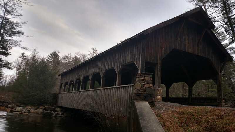 A wooden covered bridge with a steep roof, partially surrounded by trees, spans a gentle stream. The bridge features stone support pillars and large arched openings along its sides, reflecting a rustic architectural style. The scene is set against a cloudy sky, creating a serene, natural atmosphere. DuPont State Forest mountain bike trail.