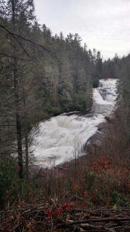 A scenic view of a waterfall cascading down rocky cliffs, surrounded by lush green forest and trees. The water flows rapidly, creating a misty spray, with some autumn foliage visible in the foreground. The sky is overcast, adding a serene, tranquil atmosphere to the landscape. DuPont State Forest mountain bike trail.