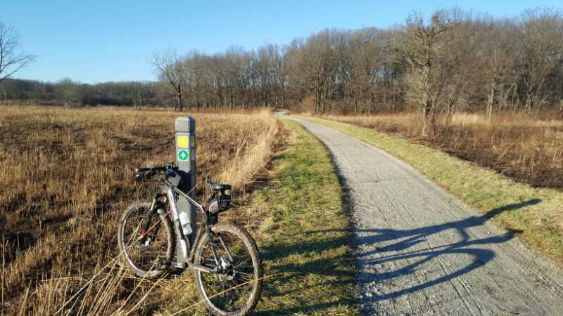 A bicycle parked next to a trail marker along a gravel pathway in a rural area. The scene features a clear blue sky, grassland, and bare trees in the background. The image captures a serene outdoor setting, ideal for cycling or walking. Palos Forest Preserve mountain bike trail.