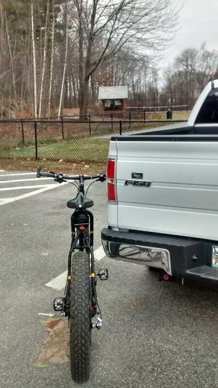 A black bicycle is parked next to a silver Ford F-150 truck in a gravel parking area. In the background, there are trees with bare branches, indicating a cool, possibly early spring or late fall setting. A wooden structure, likely a park information kiosk, is visible beyond the fence in the distance. Clifford Park mountain bike trail.